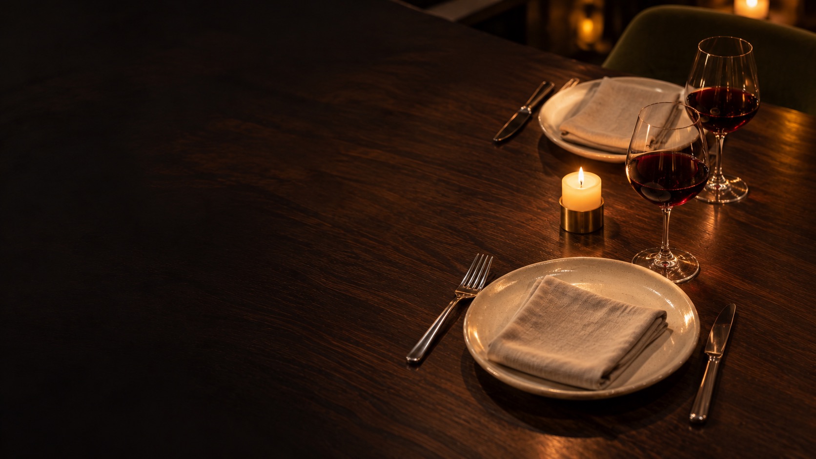 A dark timber table set for two with stoneware plates, linen napkins, two glasses of red wine and a single candle